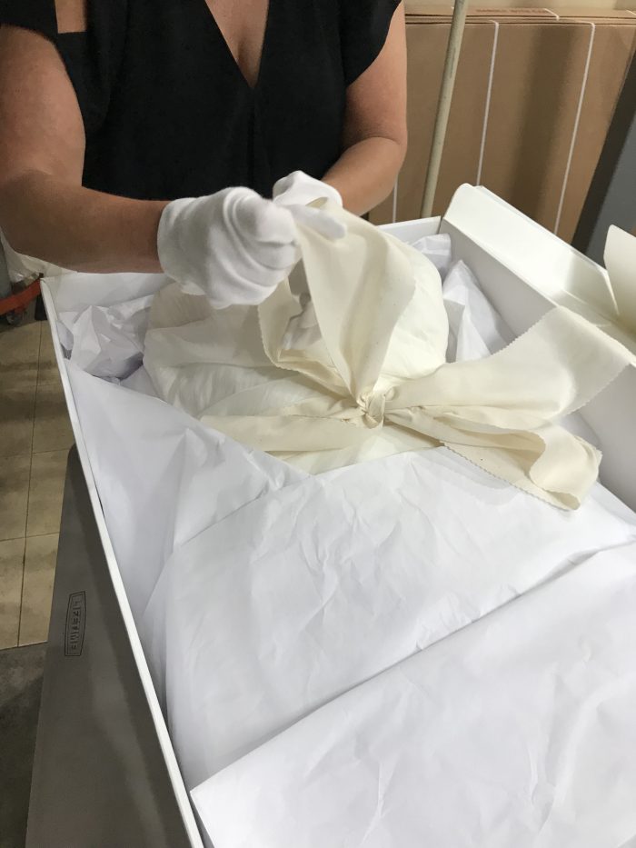A person wearing white gloves carefully unwraps a fabric bundle from a box lined with white tissue paper