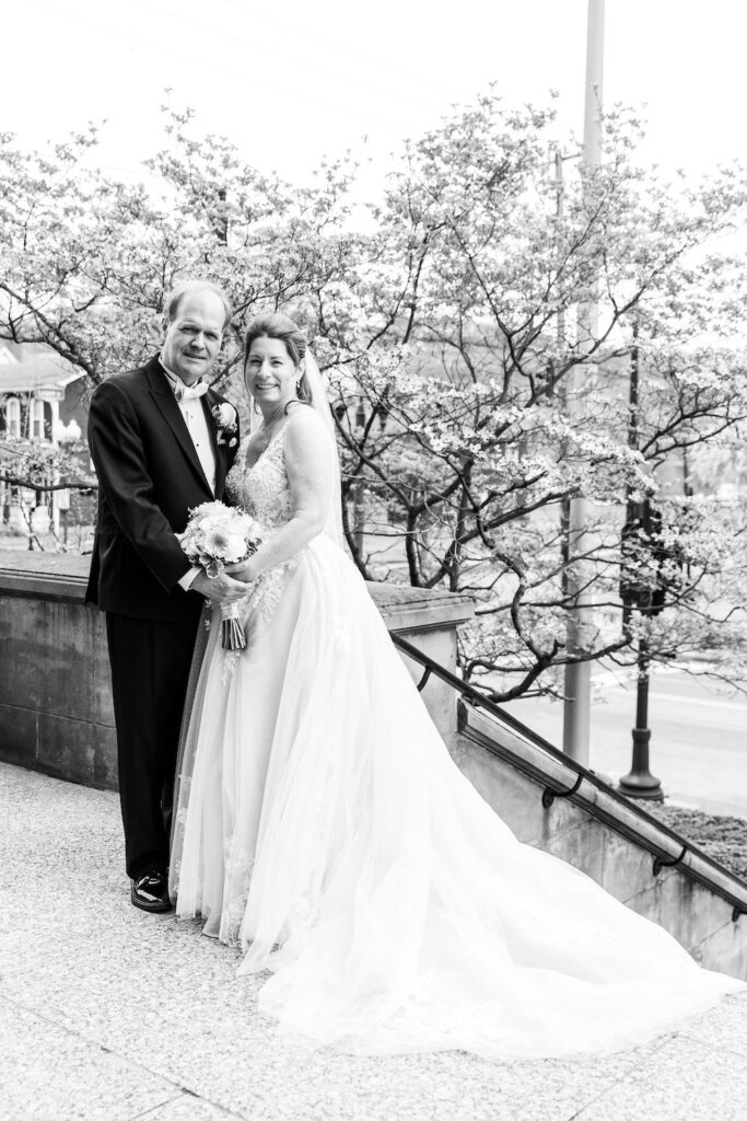 A bride and groom stand together smiling on a stone staircase, with blooming trees in the background