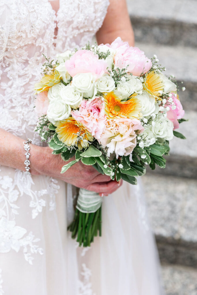 A bride holds a vibrant bouquet of white roses, pink peonies, and yellow flowers
