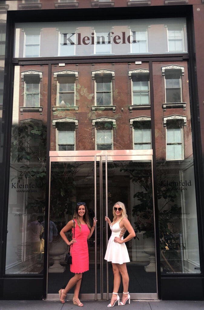 Two women stand smiling in front of the glass entrance of Kleinfeld in Manhattan