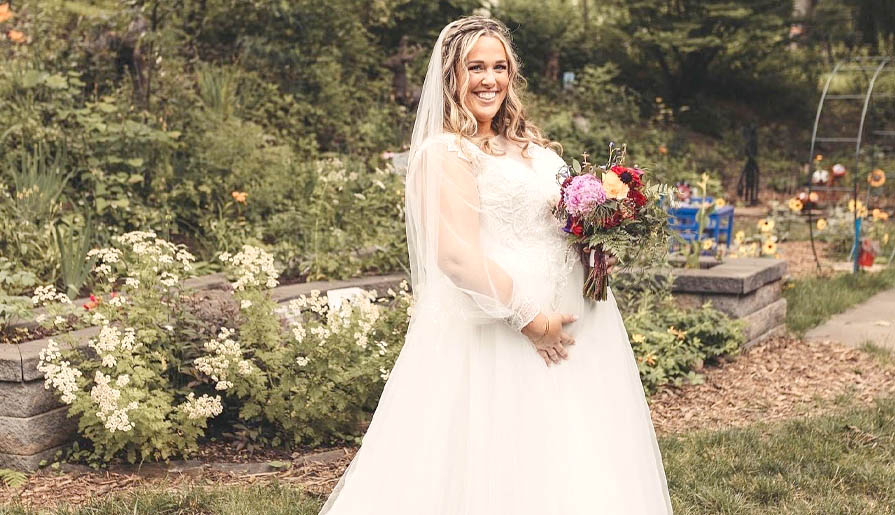 A smiling bride in a flowing white wedding dress holds a vibrant bouquet of flowers
