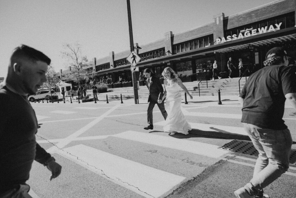 A couple in wedding attire joyfully crosses a street, holding hands