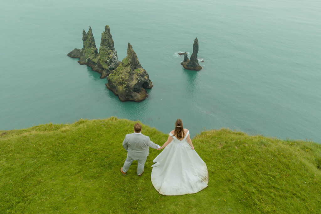 A couple stands on a grassy cliff overlooking the ocean, holding hands