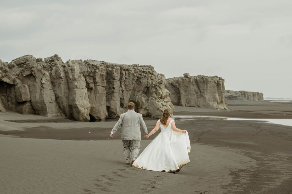 A couple in wedding attire walks hand in hand along a deserted black sand beach