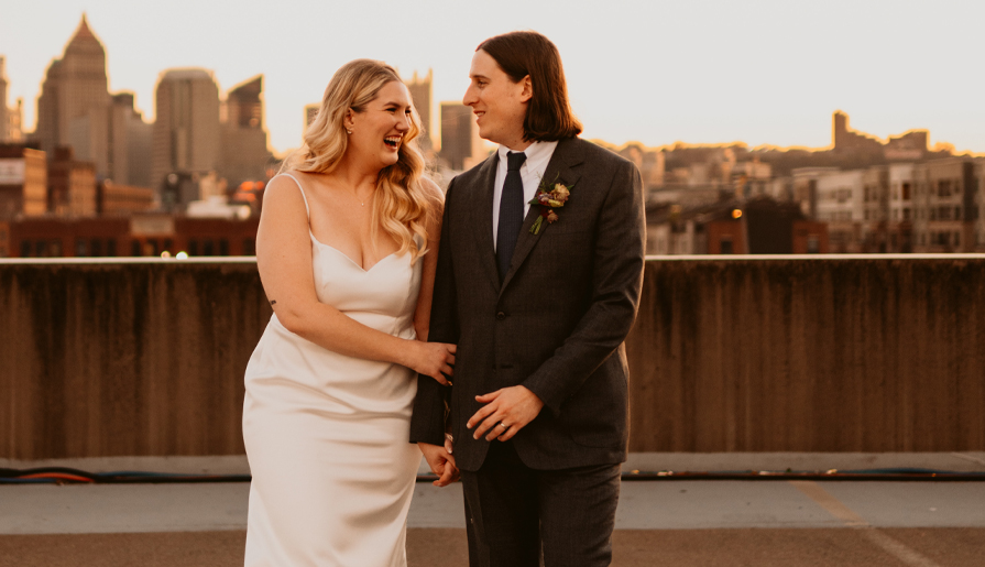 A couple is standing on a rooftop, smiling and holding hands, with a city skyline in the background