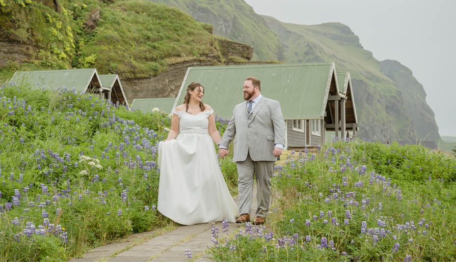A bride and groom walk hand in hand along a path lined with purple flowers