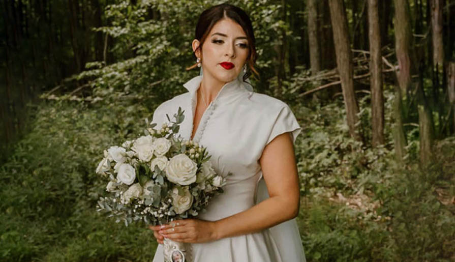 A woman in a white dress stands in a forest, holding a bouquet of white and green flowers